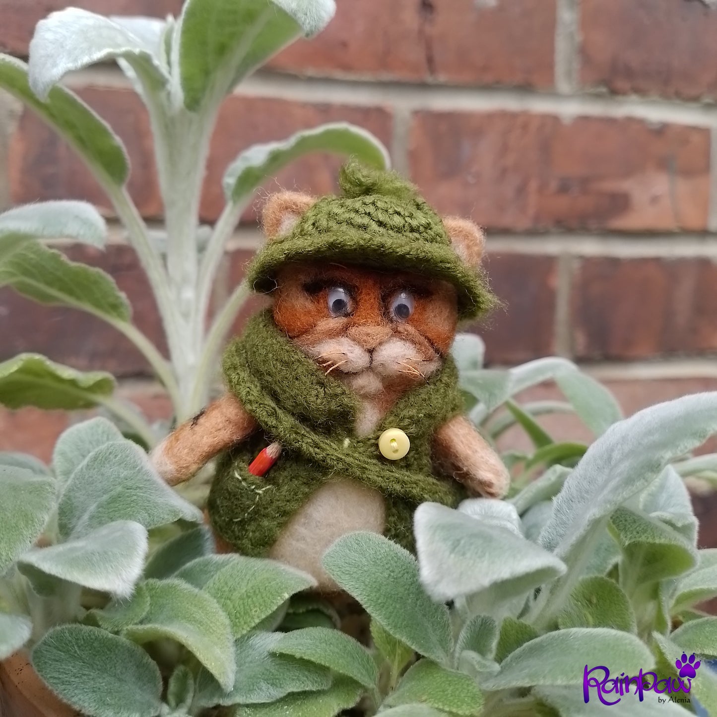 Small stuffed animal cat in a green outfit standing among plants with a brick wall background