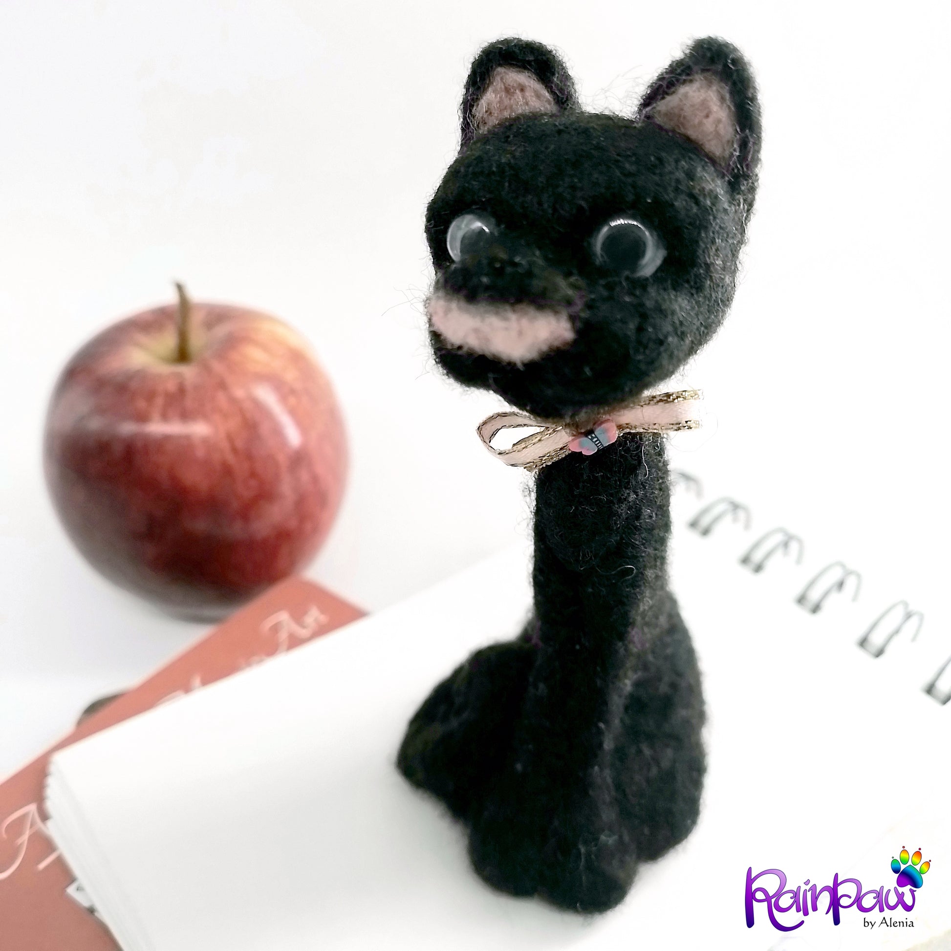 Black cat with a pink bow next to an apple on a white surface, brand 'RainPaw' visible.