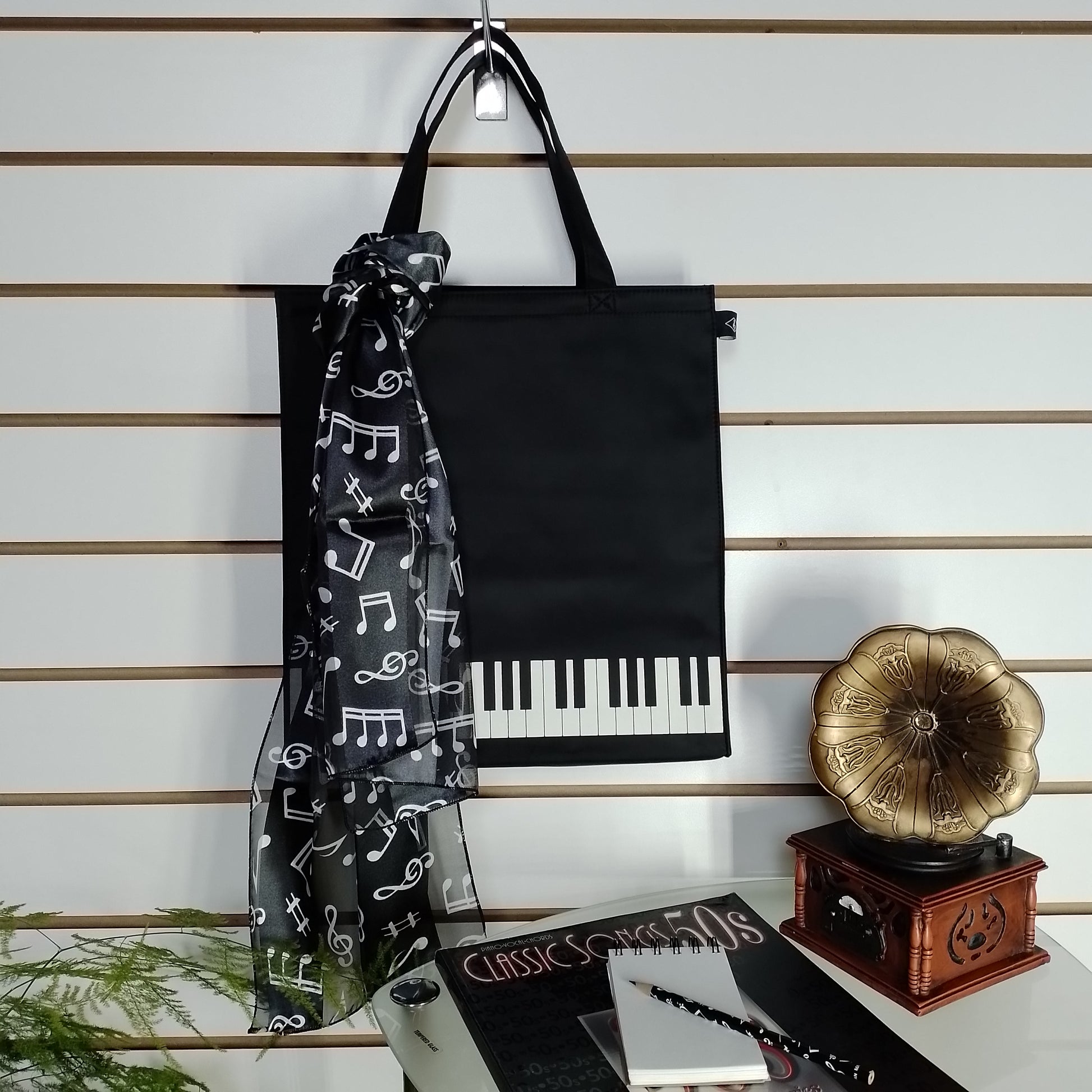 A black canvas tote bag with a piano keyboard design on the bottom half, hanging on a peg against a slatwall. A black sheer scarf with white musical notes is draped over the bag.