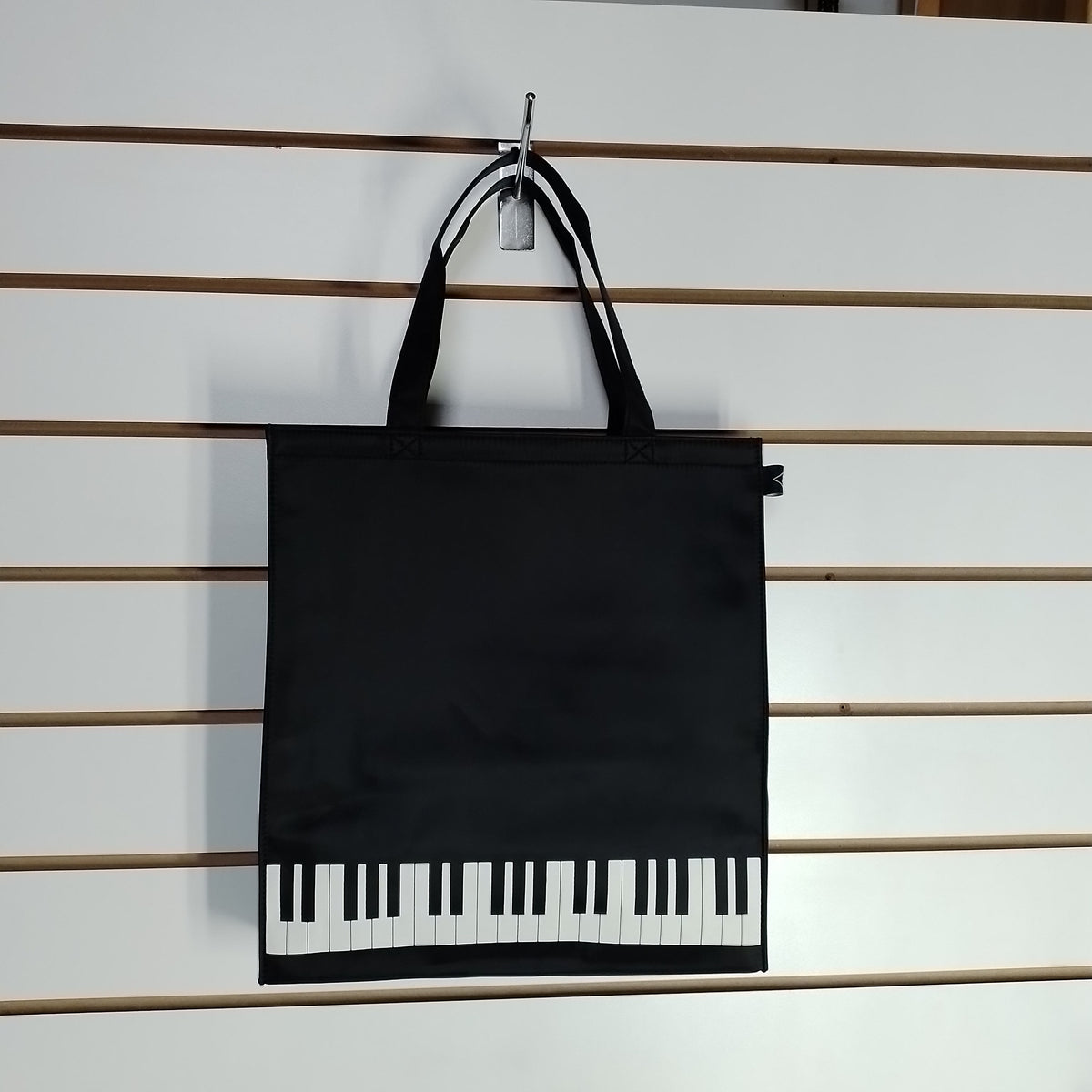 A black tote bag with piano-key graphics along the bottom, hanging from a metal hook on a white slatwall.