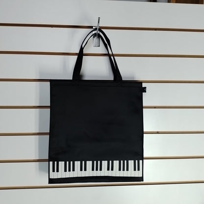 A black tote bag with piano-key graphics along the bottom, hanging from a metal hook on a white slatwall.