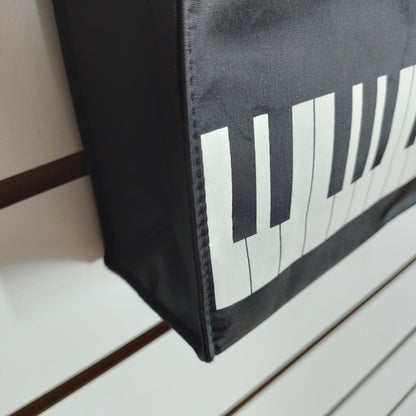 A close-up of a black fabric bag, printed with a white and black piano keyboard pattern, shown against a white slatted background.