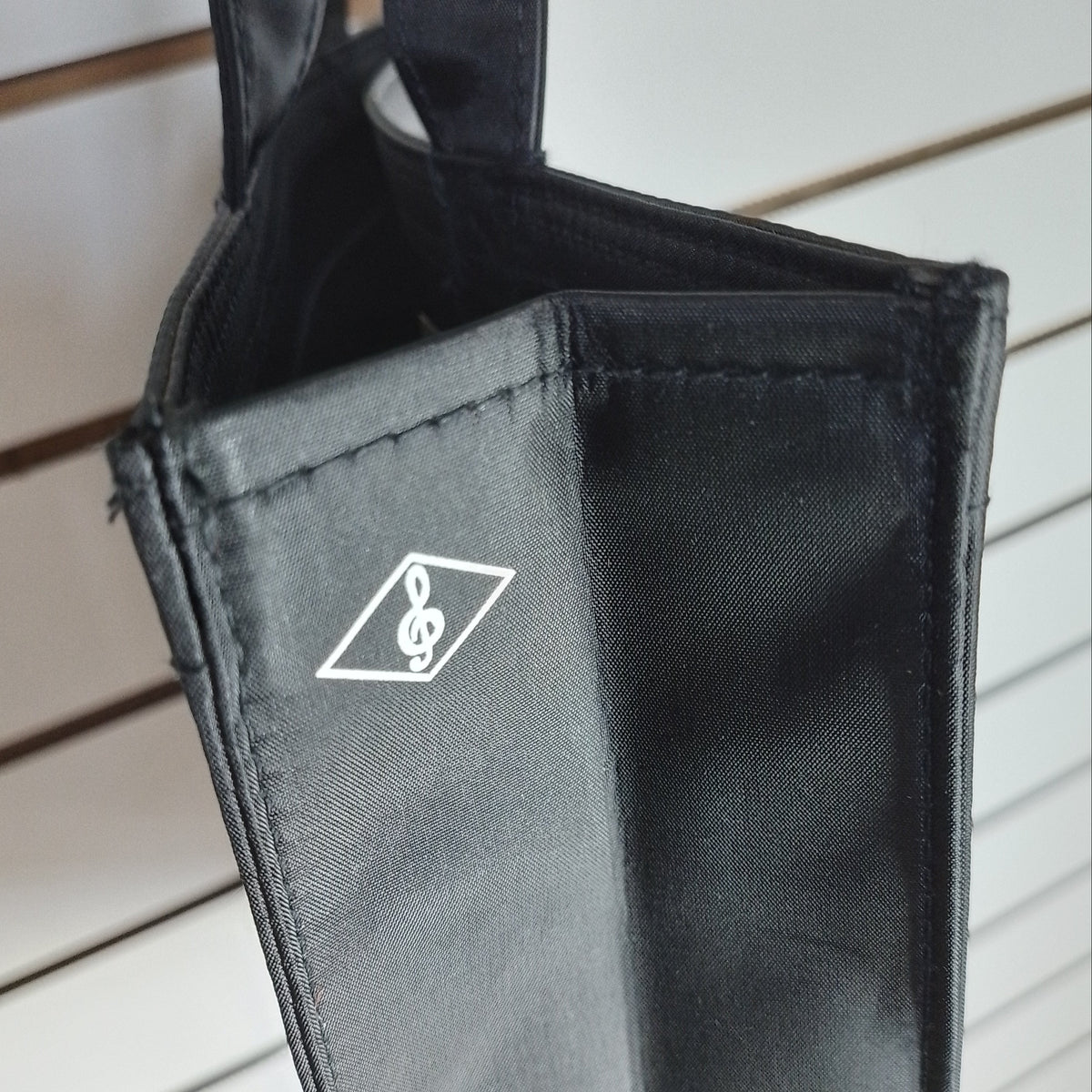 A close-up of a black fabric accessory, likely a bag or sleeve, featuring a white printed logo of a treble clef inside a diamond shape, set against a light-coloured slatted background.