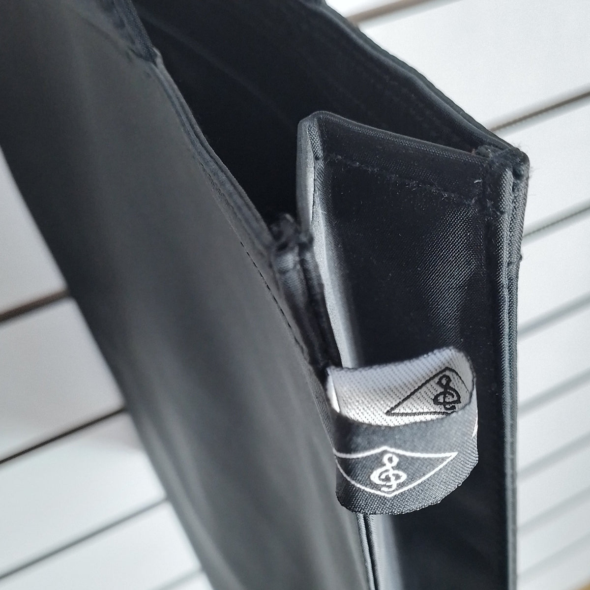 Close-up of a black fabric accessory with a small gray fabric tag featuring a white stylized logo, shown against a white slatted wall background.