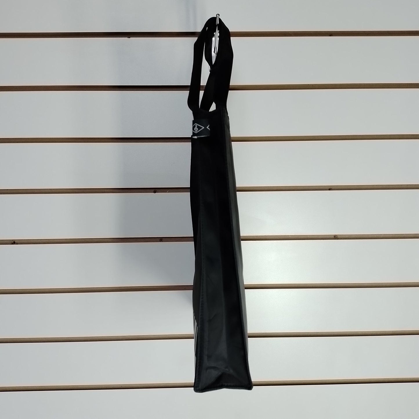 A black fabric tote bag with double handle straps hangs from a metal hook on a white slatwall.