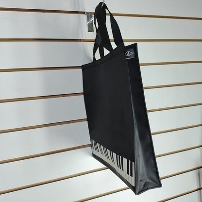 A black canvas tote bag with a white and black piano keyboard graphic along the bottom, hanging from a metal hook on a white slatwall.