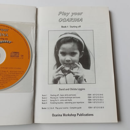 An open printed booklet or instruction sheet showing a black-and-white photo of a child and text including 'Play your Ocarina' and 'David and Dina Liggins,' with an orange CD partially visible underneath in a clear sleeve.