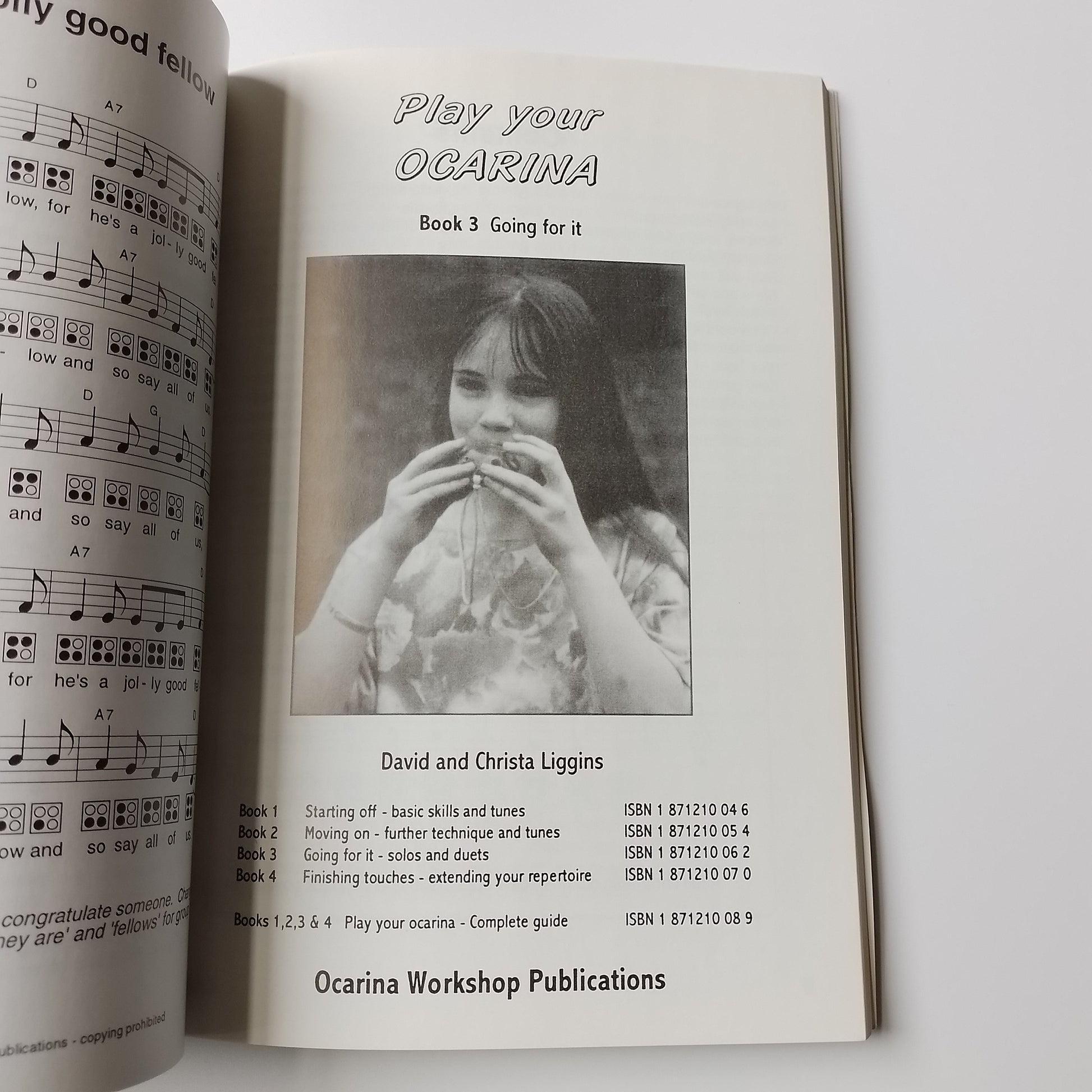 An open book or booklet showing printed sheet music and a black-and-white photo of a person holding an ocarina. The visible text includes 'Play your Ocarina', 'David and Christa Liggins', and 'Ocarina Workshop Publications'.