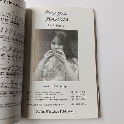 An open book or booklet showing printed sheet music and a black-and-white photo of a person holding an ocarina. The visible text includes 'Play your Ocarina', 'David and Christa Liggins', and 'Ocarina Workshop Publications'.