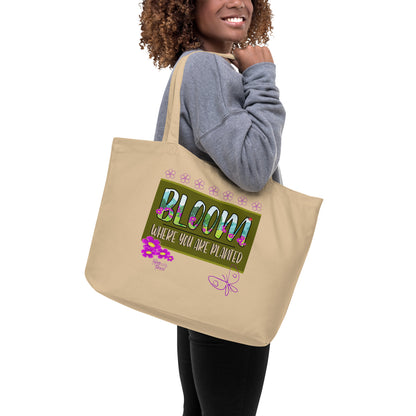 Person holding a beige tote bag with 'Bloom Where You Are Planted' design.