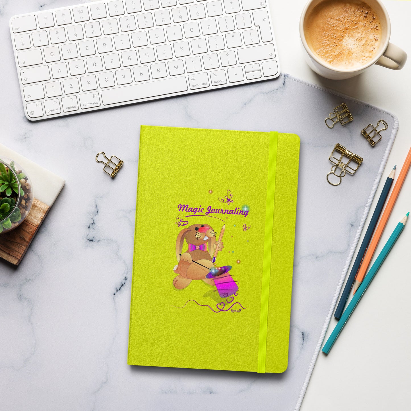 A lime-green journal with a rubber band lies on a marble desk alongside a keyboard, coffee, and stationery. The cover features a bunny holding a magic wand and a pencil pulled from its top hat, surrounded by butterflies, colourful stars, and hearts, giving the journal an enchanting feel. The words "Magic Journaling" are written on the cover.