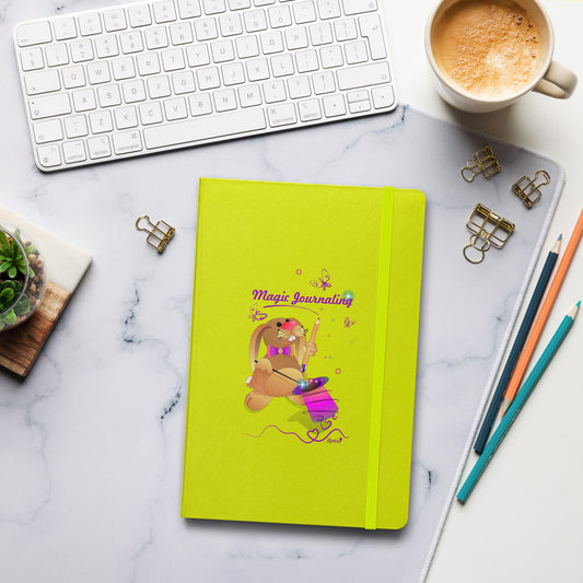A lime-green journal with a rubber band lies on a marble desk alongside a keyboard, coffee, and stationery. The cover features a bunny holding a magic wand and a pencil pulled from its top hat, surrounded by butterflies, colourful stars, and hearts, giving the journal an enchanting feel. The words "Magic Journaling" are written on the cover.