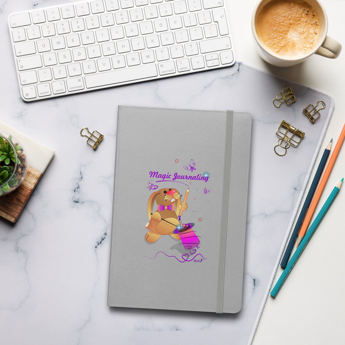 A silver journal with a rubber band lies on a marble desk alongside a keyboard, coffee, and stationery. The cover features a bunny holding a magic wand and a pencil pulled from its top hat, surrounded by butterflies, colourful stars, and hearts, giving the journal an enchanting feel. The words "Magic Journaling" are written on the cover.