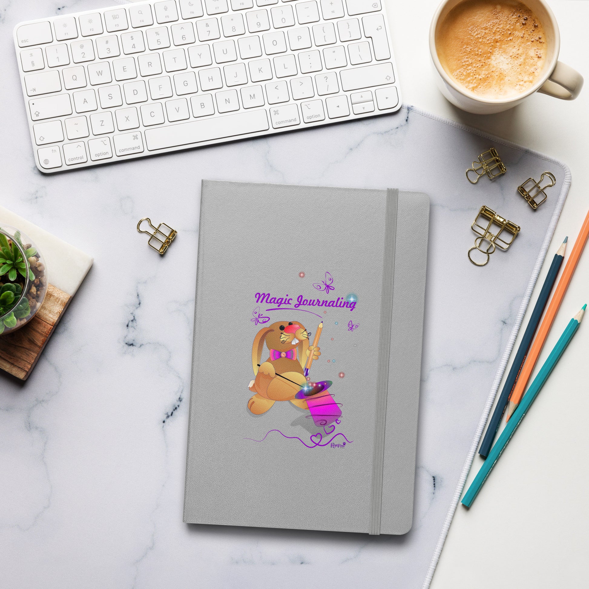 A silver journal with a rubber band lies on a marble desk alongside a keyboard, coffee, and stationery. The cover features a bunny holding a magic wand and a pencil pulled from its top hat, surrounded by butterflies, colourful stars, and hearts, giving the journal an enchanting feel. The words "Magic Journaling" are written on the cover.