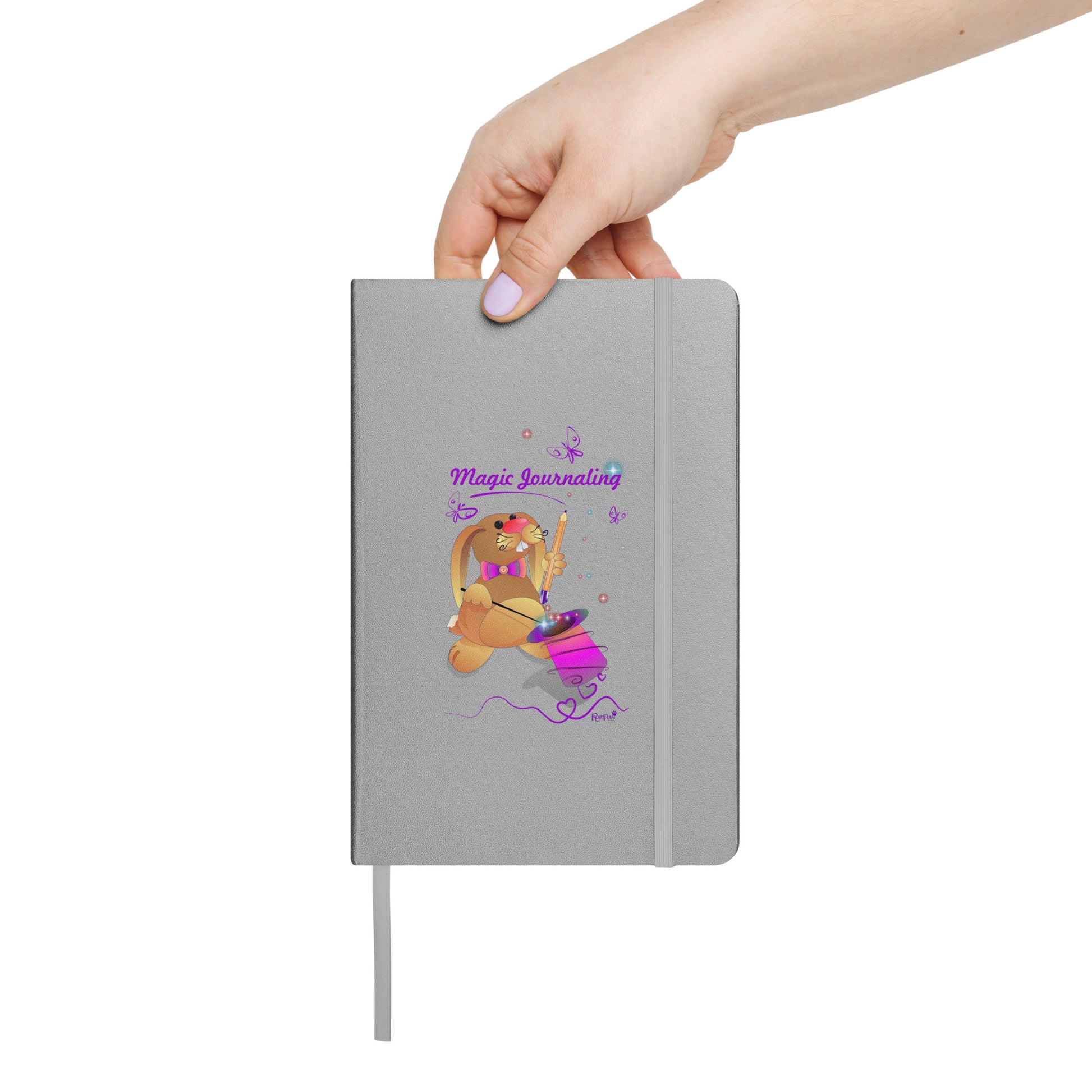 Silver notebook held by a hand on a white background. - The cover features a bunny holding a magic wand and a pencil pulled from its top hat, surrounded by butterflies, colourful stars, and hearts, giving the journal an enchanting feel. The words "Magic Journaling" are written on the cover.