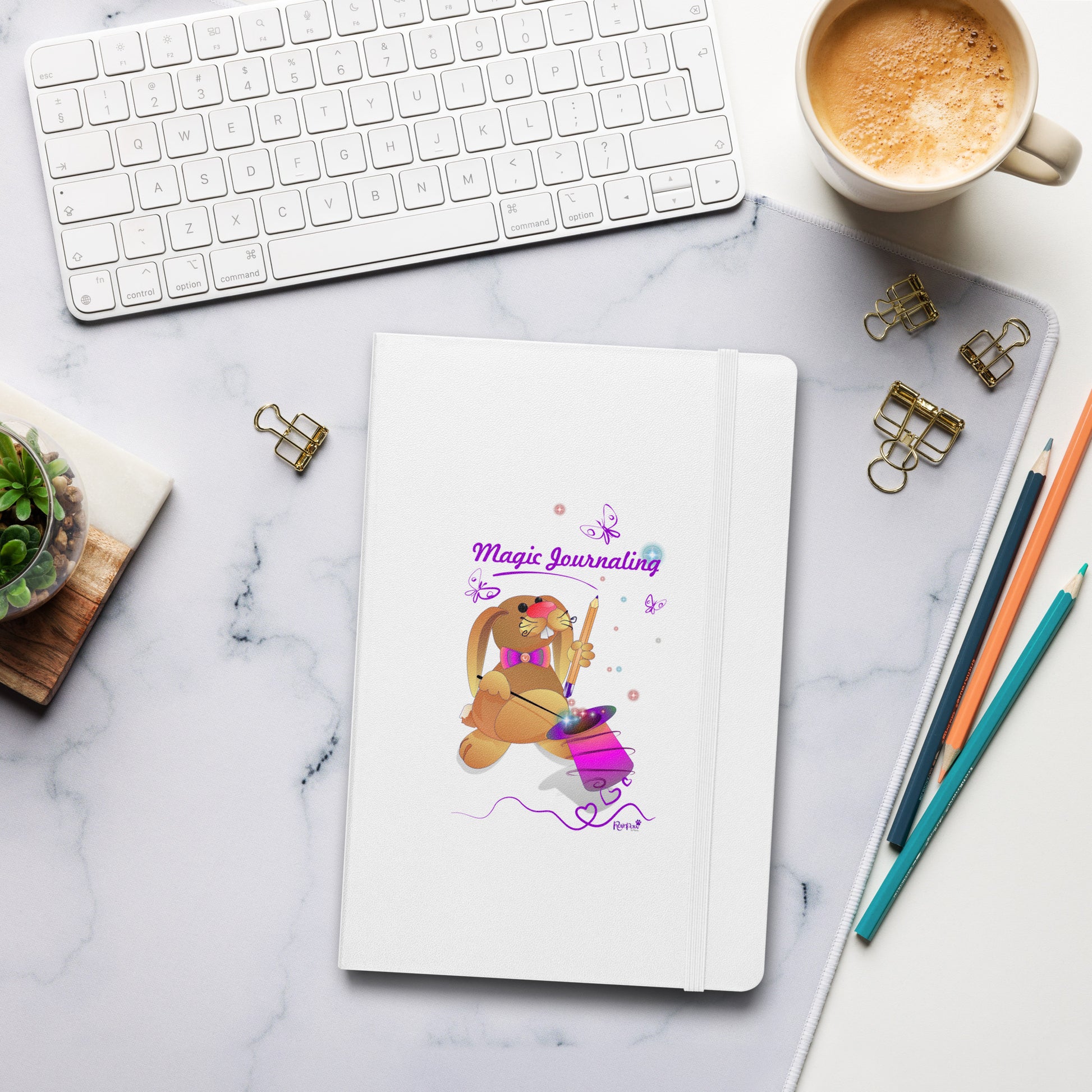 A white journal with a rubber band lies on a marble desk alongside a keyboard, coffee, and stationery. The cover features a bunny holding a magic wand and a pencil pulled from its top hat, surrounded by butterflies, colourful stars, and hearts, giving the journal an enchanting feel. The words "Magic Journaling" are written on the cover.