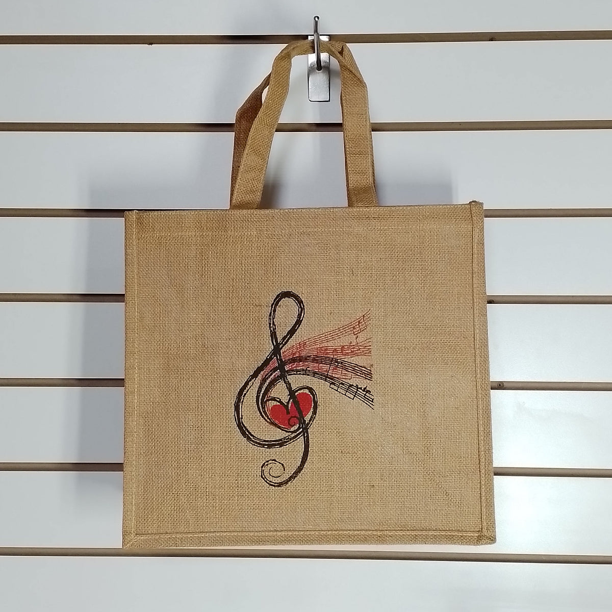 A natural jute tote bag with a treble clef and red heart graphic, hanging from a metal hook on a white slatwall.
