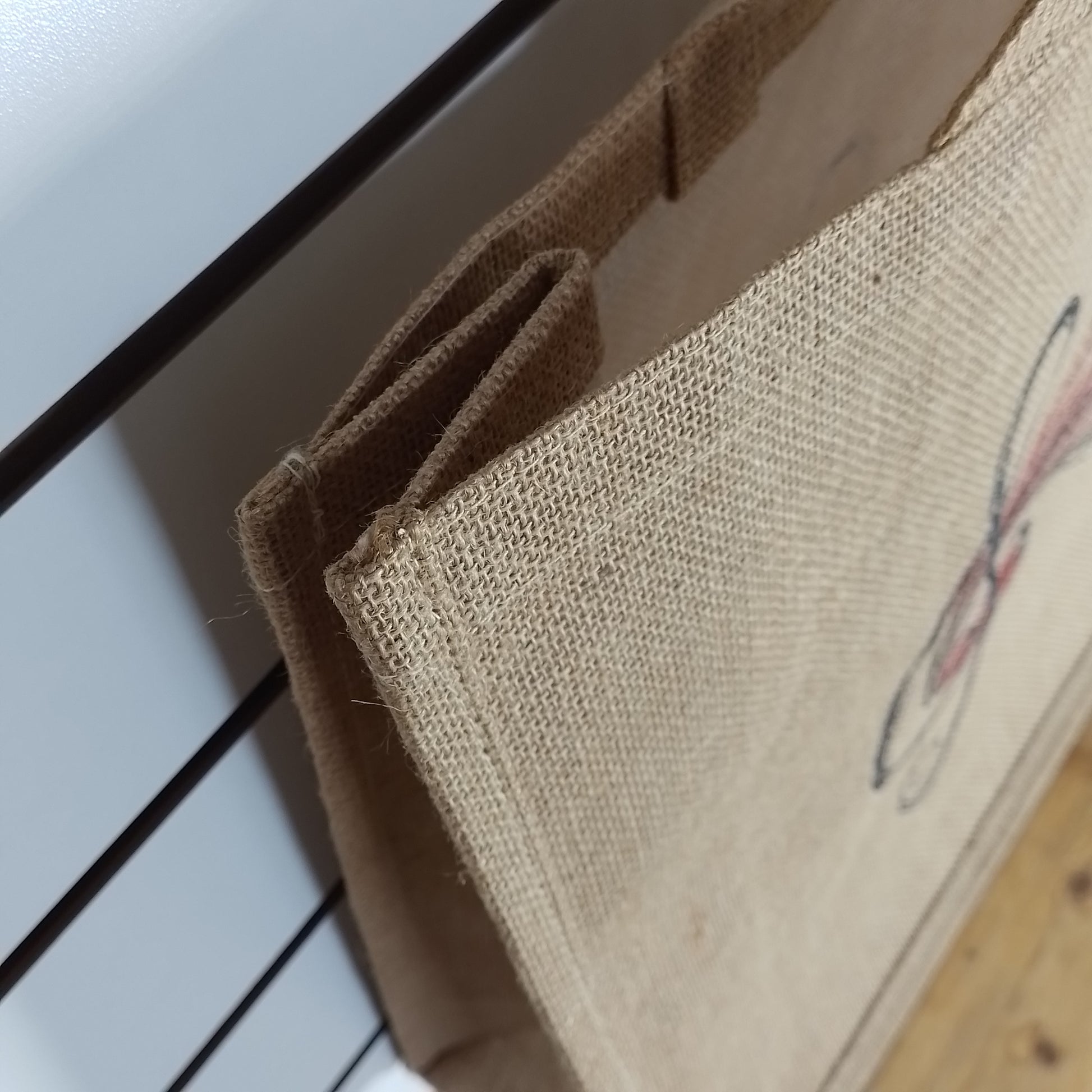 Close-up of a natural burlap tote bag with brown handles, leaning against a white slatted surface on a light wood floor. A partial graphic is visible on the bag.