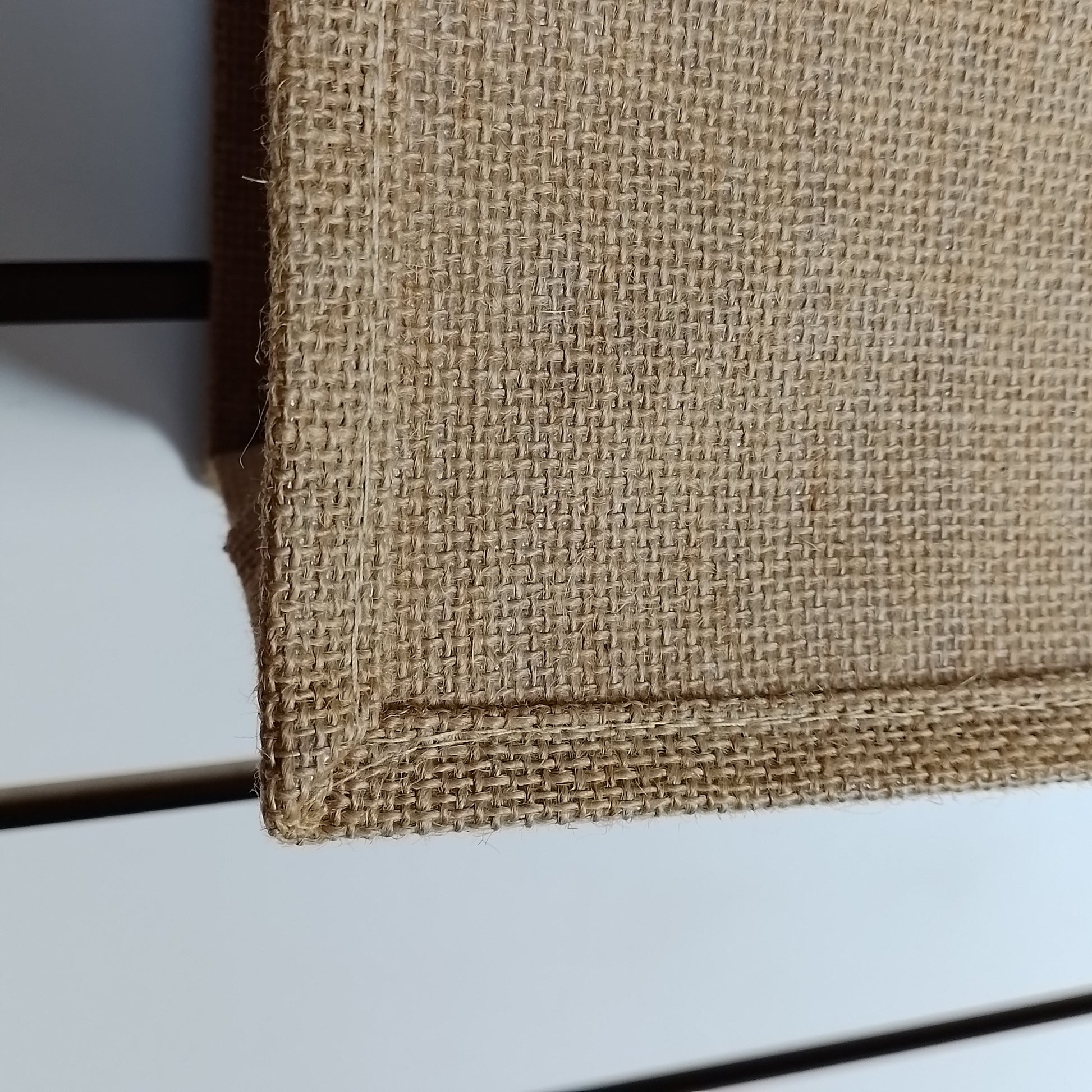 Close-up of a natural jute or burlap fabric surface with a visible woven texture and stitched edge, shown against a light neutral background.