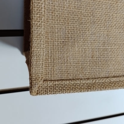 Close-up of a natural jute or burlap fabric surface with a visible woven texture and stitched edge, shown against a light neutral background.