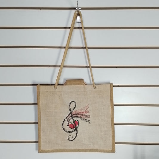 A natural jute tote bag with tan shoulder straps, featuring a black treble clef and red heart graphic with pink ribbon details, hanging on a white slatwall.