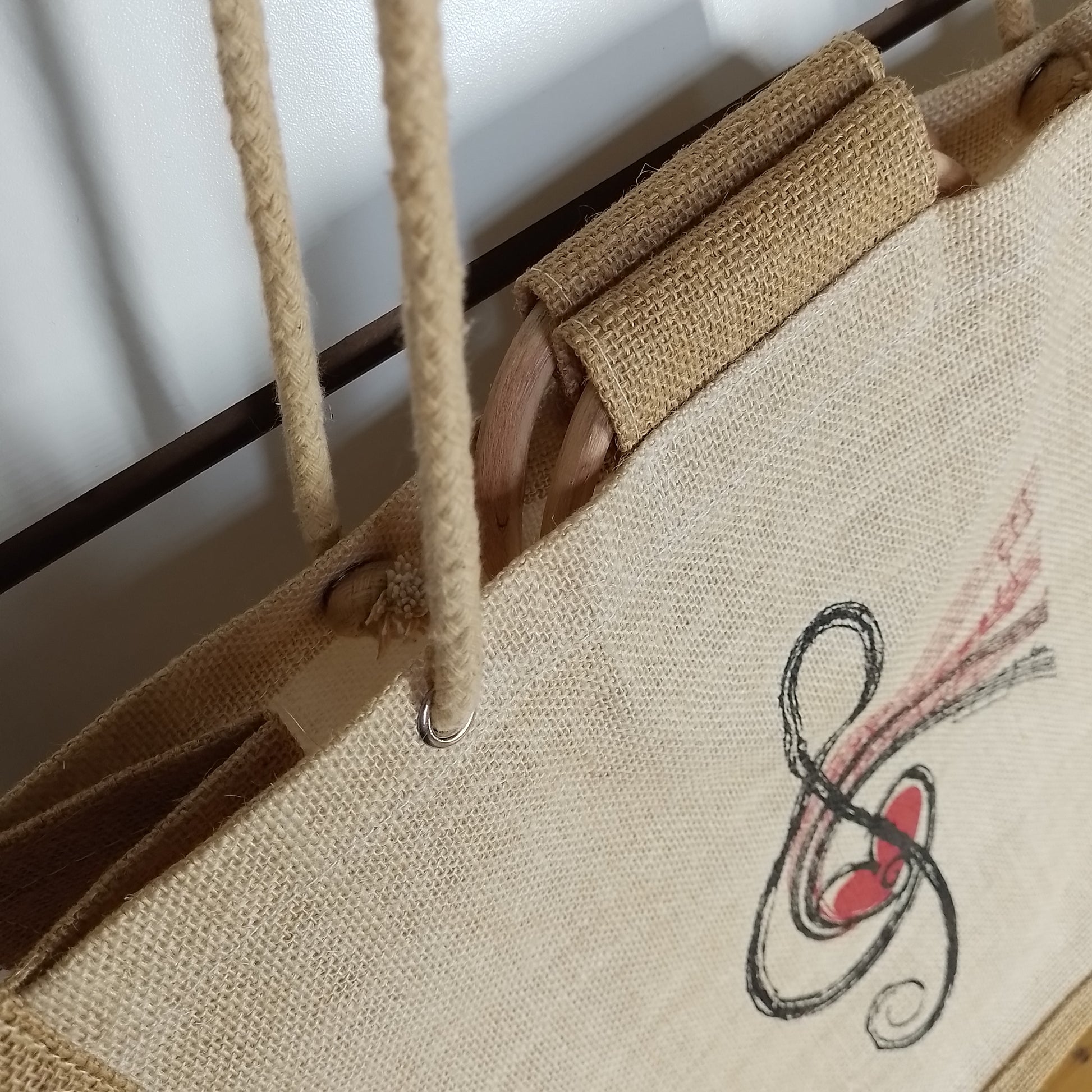 Close-up of a natural jute tote bag with rope handles, a wooden handle insert, and a black treble clef graphic with a red heart on the front.
