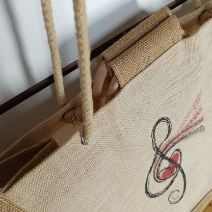 Close-up of a natural jute tote bag with rope handles, a wooden handle insert, and a black treble clef graphic with a red heart on the front.