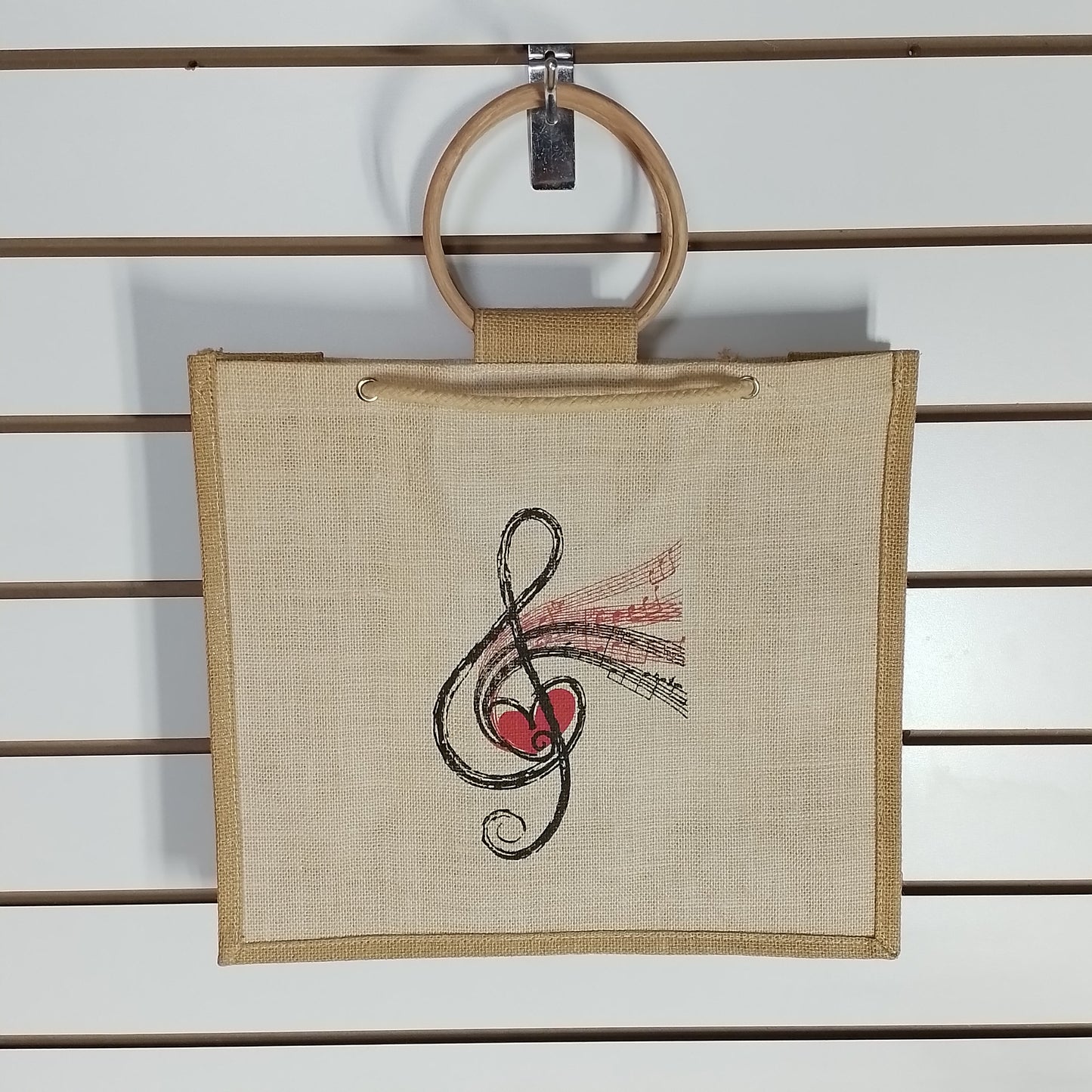 A square beige canvas tote bag with a wooden ring handle, hanging on a white slatwall. The bag features a black treble clef and red heart graphic with pink musical notes printed on the front.