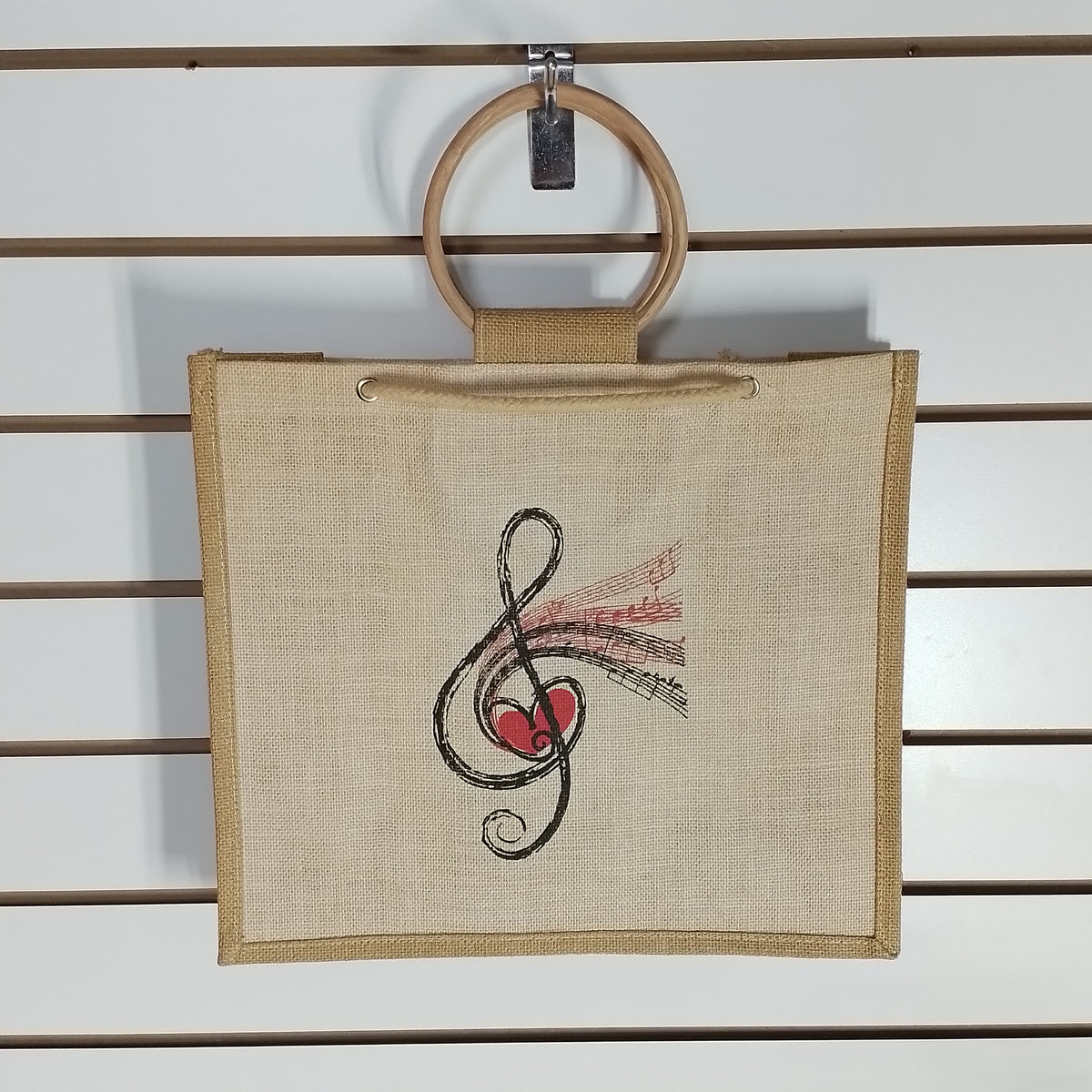 A square beige canvas tote bag with a wooden ring handle, hanging on a white slatwall. The bag features a black treble clef and red heart graphic with pink musical notes printed on the front.