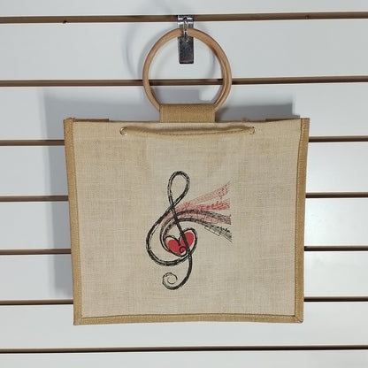 A square beige canvas tote bag with a wooden ring handle, hanging on a white slatwall. The bag features a black treble clef and red heart graphic with pink musical notes printed on the front.