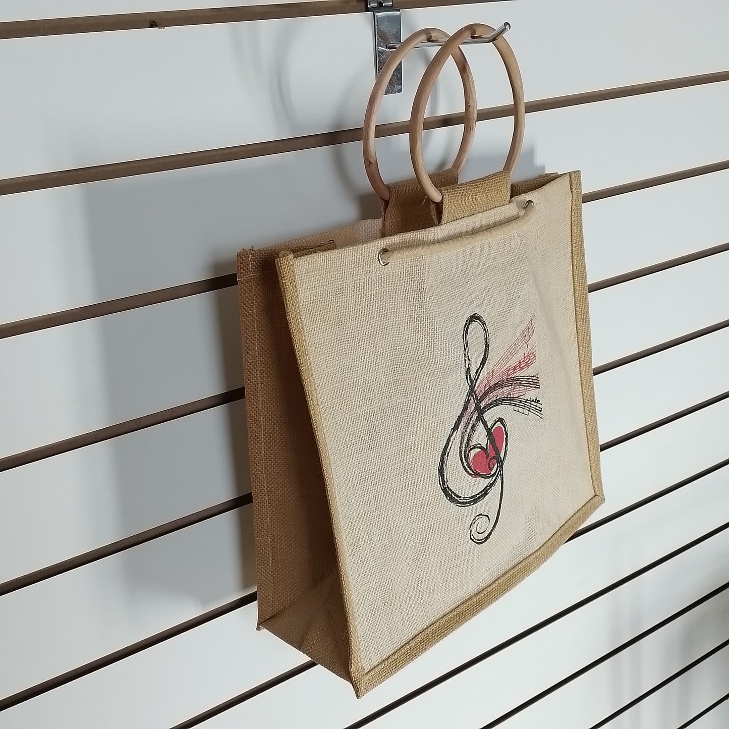 A natural jute tote bag with double wooden handles, featuring a black and red musical treble clef with a heart design on the front, hanging on a brown slatwall display.