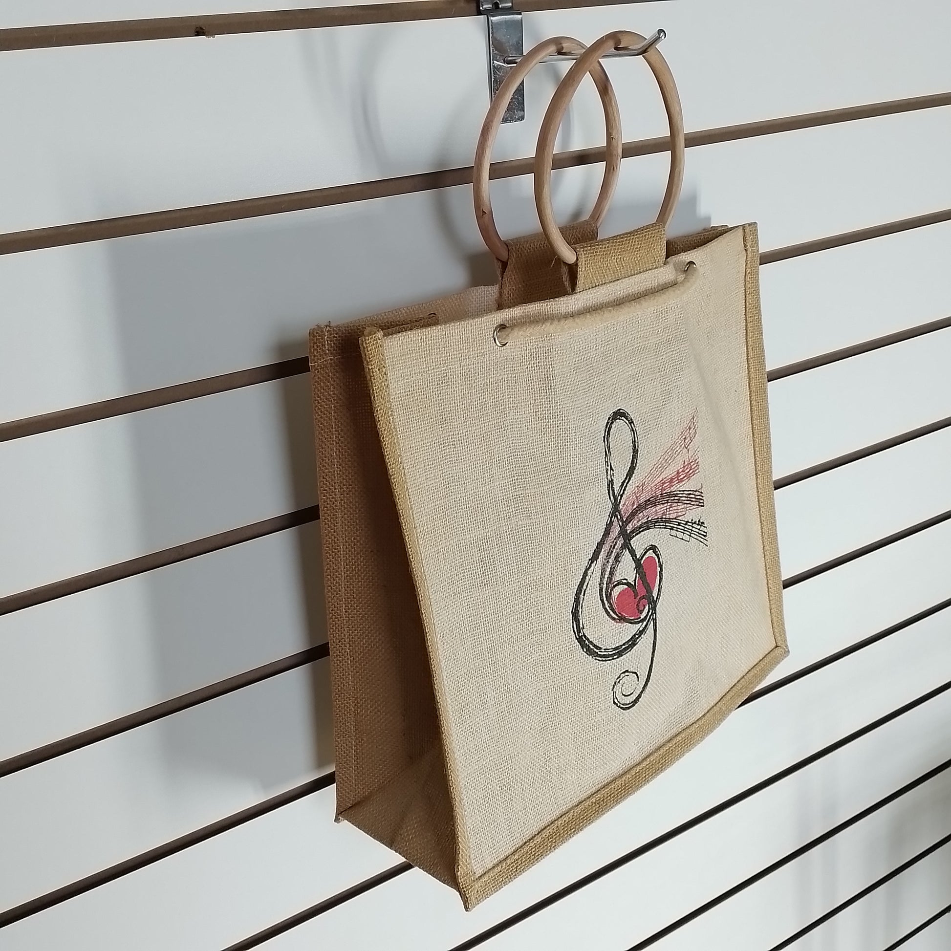 A natural jute tote bag with double wooden handles, featuring a black and red musical treble clef with a heart design on the front, hanging on a brown slatwall display.