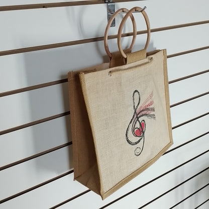 A natural jute tote bag with double wooden handles, featuring a black and red musical treble clef with a heart design on the front, hanging on a brown slatwall display.
