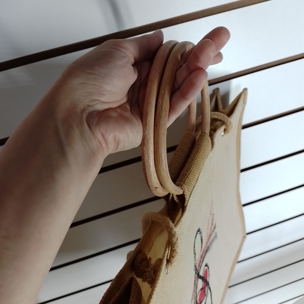 A close-up of a hand holding the double wooden handles of a beige canvas tote bag with a small red and black graphic print on the side, set against a white slatted background.