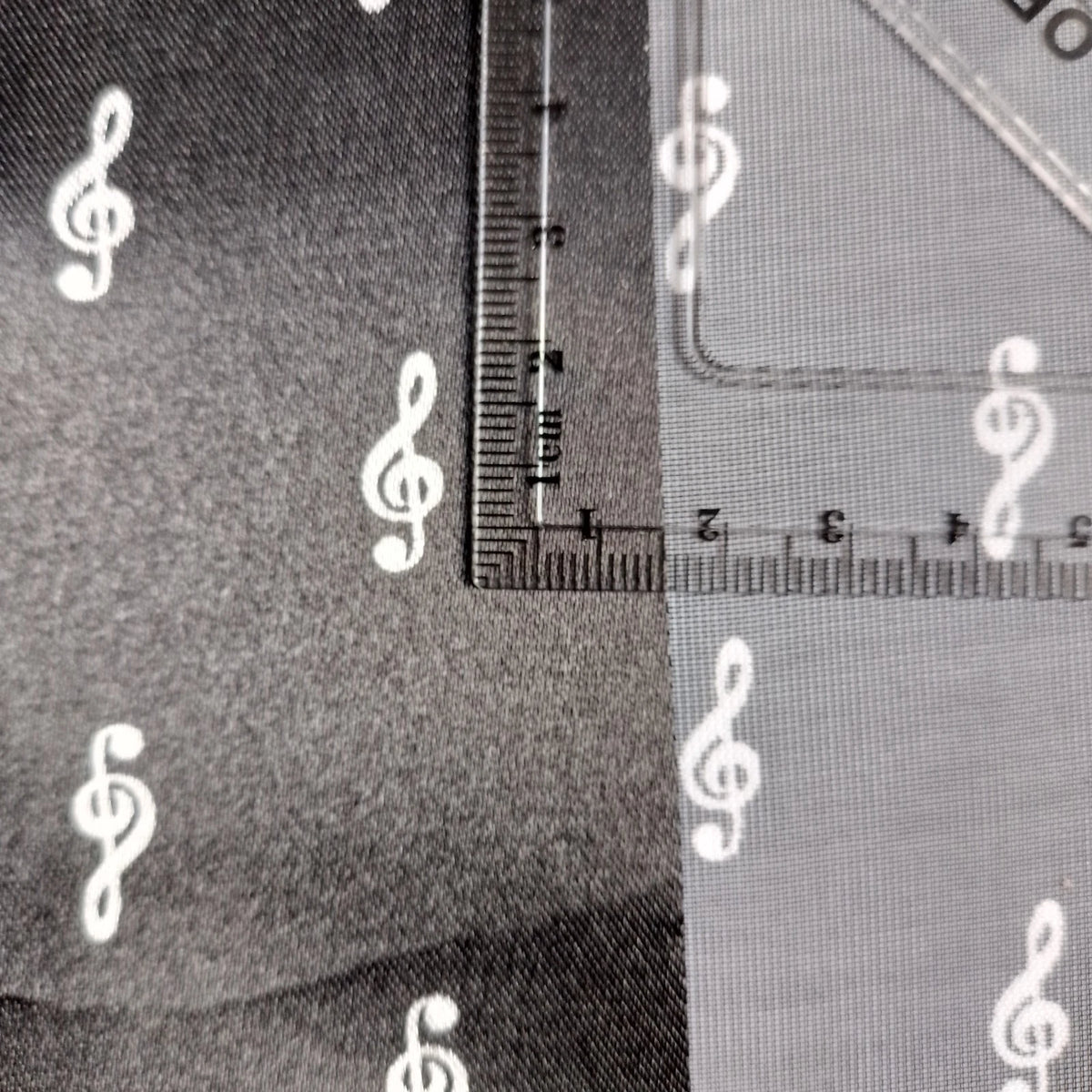 Close-up of a fabric or material sample with white repeating white treble clef pattern, shown alongside a clear ruler for scale.