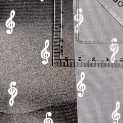 Close-up of a fabric or material sample with white repeating white treble clef pattern, shown alongside a clear ruler for scale.
