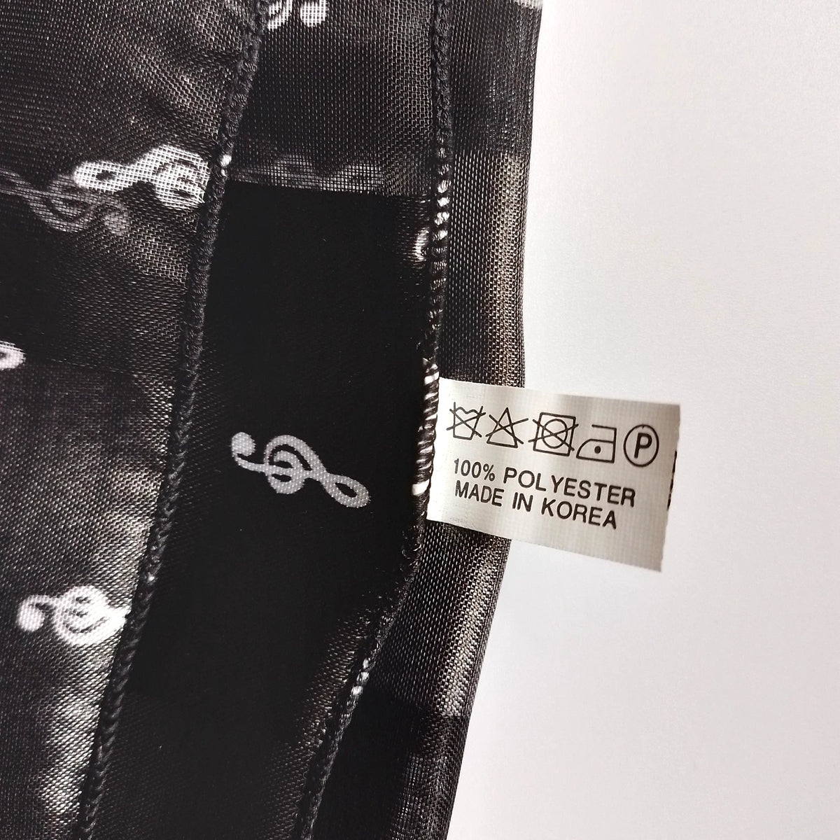 Close-up of a black patterned fabric with white treble clefs and a sewn-on care label reading 100% POLYESTER MADE IN KOREA.