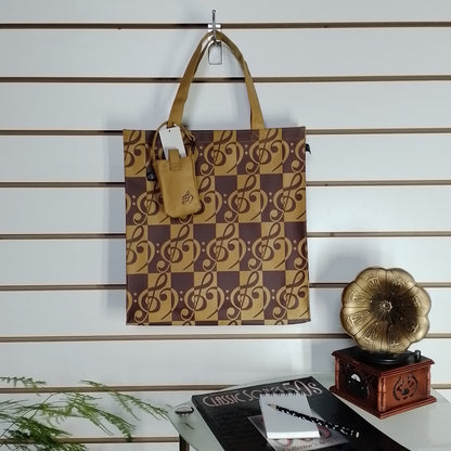 A brown and tan checkered tote bag with treble clefs hanging on a wall hook, with a small matching pouch attached. The background is a white slatwall with decorative props including a gramophone and a plant.