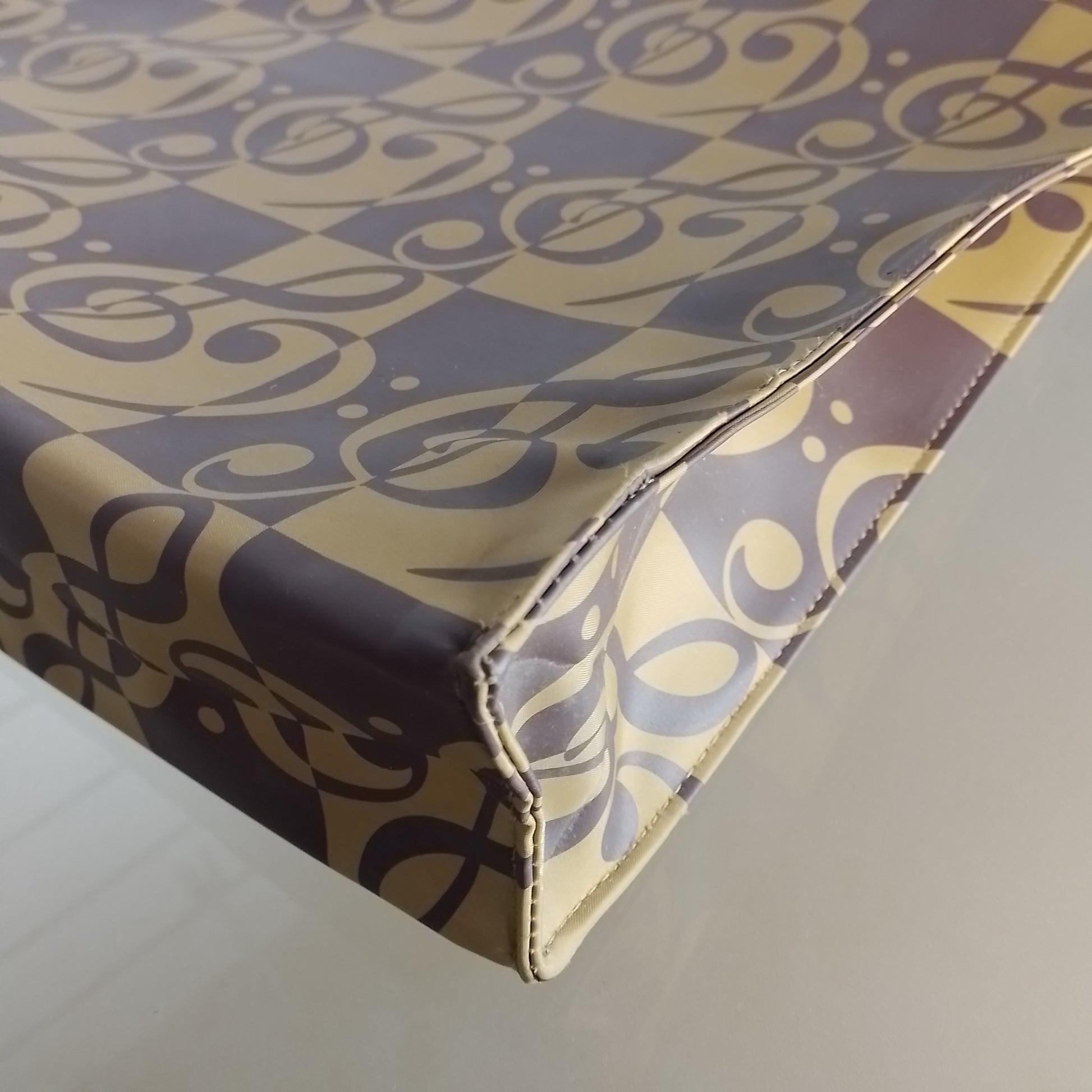 A close-up of a corner of a soft-sided bag or case with a repeating scroll pattern in dark brown and beige, resting on a light gray surface.