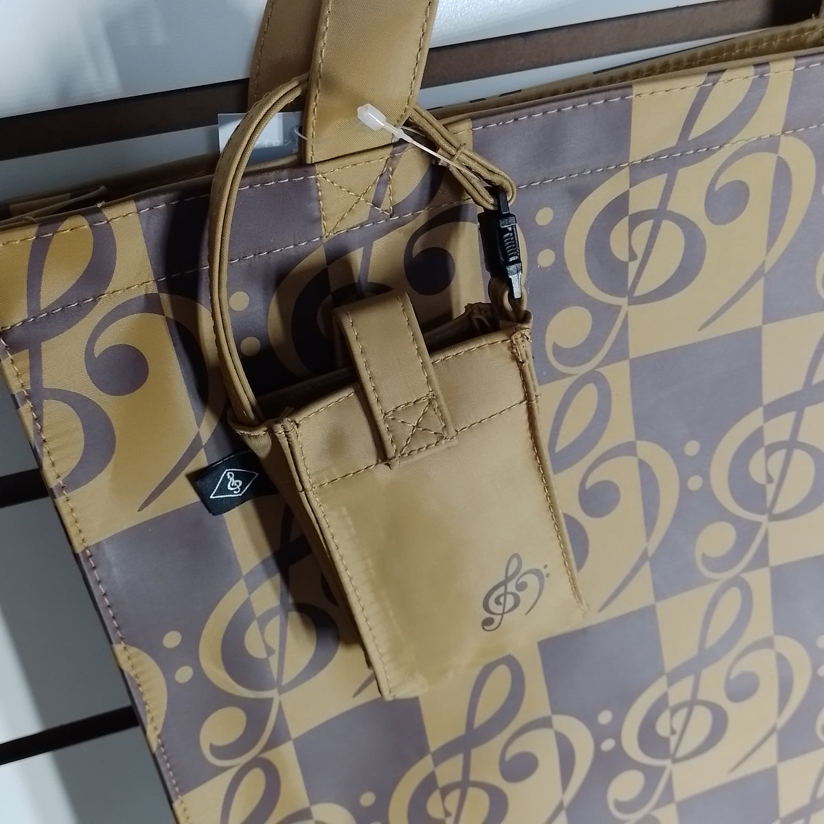 A tan nylon accessory pouch with a small treble clef logo, attached to a matching patterned tote bag with a brown and tan checkered design and musical notes.