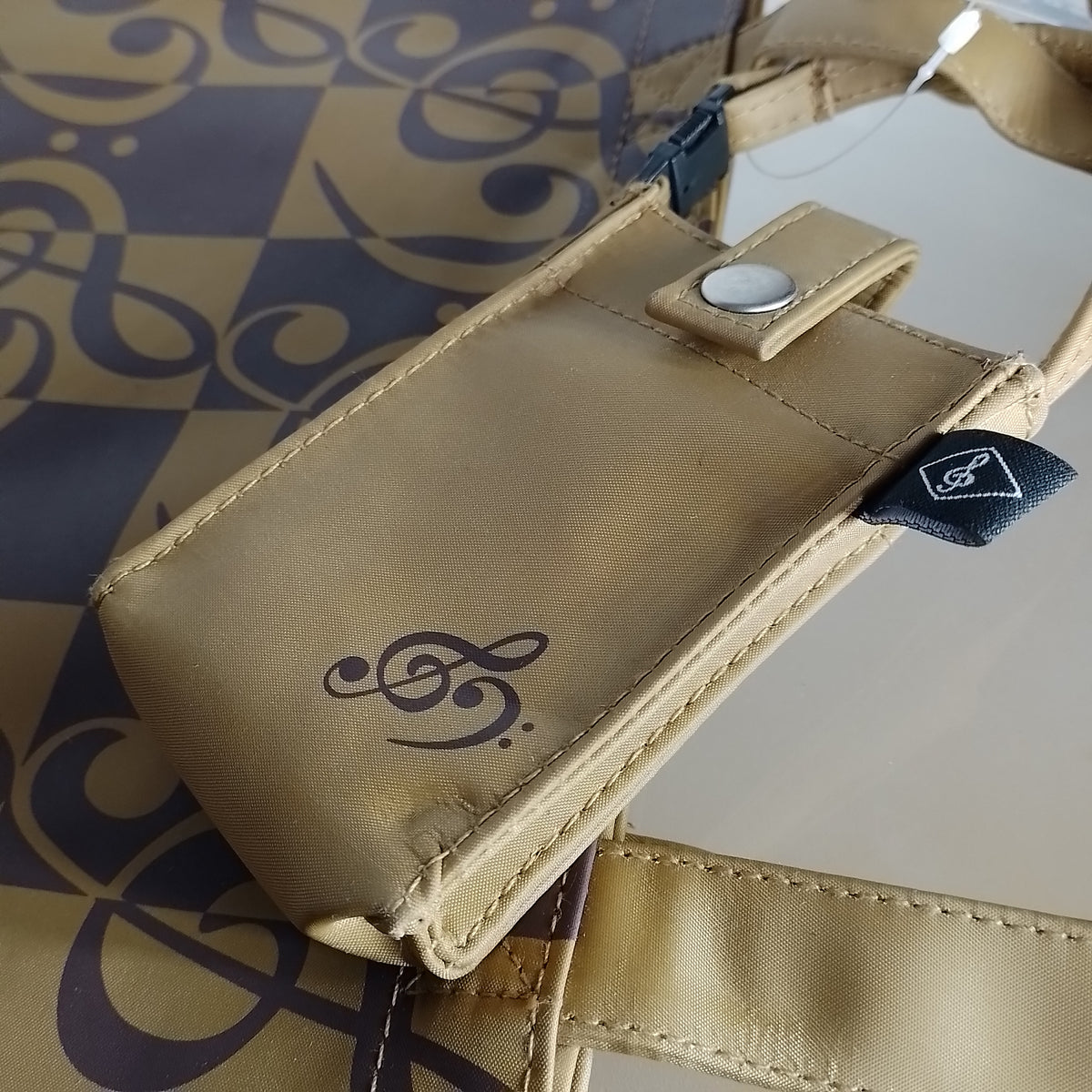A beige  phone pouch with a treble and bass clef logo, featuring a snap-button closure and a black brand tag, resting on a patterned bag.